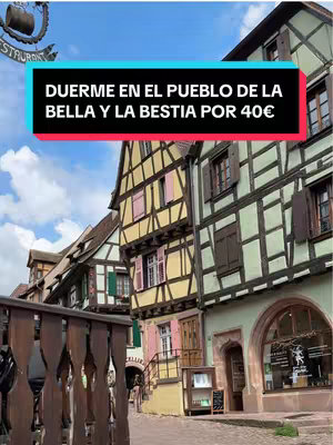 The image shows a street view of a town composed of colorful historic buildings. The architectural style appears European, with timber-framed structures and vibrant hues. Above, a text block in a light blue rectangle reads: "DUERME EN EL PUEBLO DE LA BELLA Y LA BESTIA POR 40€".