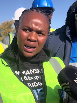 A woman with dark hair in braids, wearing a lime green safety vest over a black hooded sweatshirt, looks directly into the camera with a concerned expression. Behind her, blurred figures wear blue and white hard hats. The background suggests an outdoor environment, possibly a construction or industrial site. A dark microphone is visible to the right of the frame.