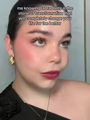 A close-up shot of a person with light skin and dark hair, framed from the chest up. The person looks towards the camera with eyes slightly upwards, lips are glossy red, and cheeks are blushed. The background is a plain, light grey or white. The person wears a black, off-the-shoulder top and gold-colored earrings.