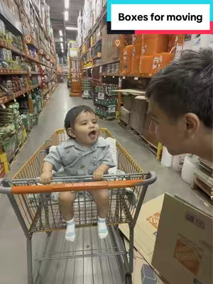 A candid shot inside a Home Depot-like store. A young child is seated in a shopping cart being pushed down an aisle. The child is looking and speaking towards another person not visible in the frame. The aisle is filled with tall shelves stocked with various items.