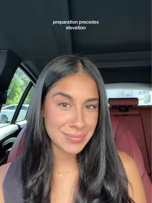 A selfie-style video taken from inside a car. The woman is smiling, and the focus is on her face. The background shows the inside of the car and the outside view through the window..