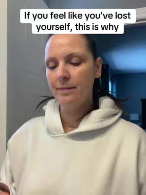 A woman in a white hoodie is centered in the frame and looking slightly downward with a neutral expression. The top caption text reads "If you feel like you've lost yourself, this is why". The background is a plain wall and a door.