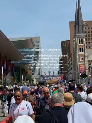A crowd of people walking down a street towards the camera on a bright, sunny day, buildings and a church with a tall, pointed spire in the background. Text overlay is partially visible.