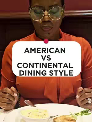 A woman wearing large glasses and an orange top is centered in the frame, with a white oval containing the text 'AMERICAN VS CONTINENTAL DINING STYLE' in bold black letters in the center. Her hands are near a plate of food and a spoon on the table with more food items.