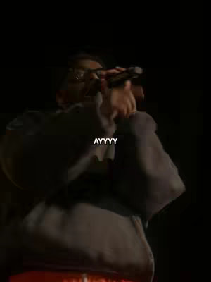 The image showcases a person, seemingly on stage, holding a microphone and singing. The person is dressed in a dark hoodie and glasses, with only their upper body visible against a dark background. The word "AYYYY" is overlaid in white font.