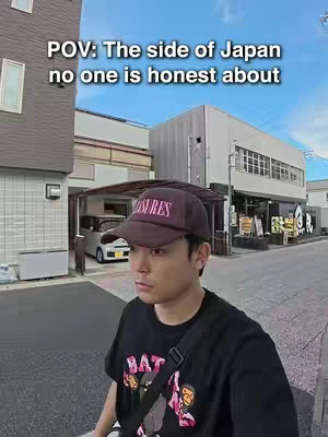 A man wearing a cap and a black t-shirt stands on a street, looking slightly to the side. He's in front of buildings typical of a Japanese town with a bright sky in the background.