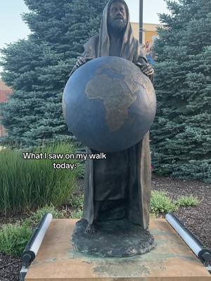 The cover image shows a bronze statue of a man holding a large globe in front of a backdrop of green trees. The statue is placed on a stone base and flanked by what seem to be two light fixtures. Text at the bottom of the image reads 'What I saw on my walk today:'. The color palette uses natural tones of greens, browns, and the bronze of the statue.