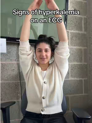 A woman stands with her arms raised overhead, her hands clasped. She is wearing a light-colored cardigan over a white top. The background consists of a grey brick wall, and a dark TV screen. The video has text overlayed that says "Signs of hyperkalemia on an ECG".