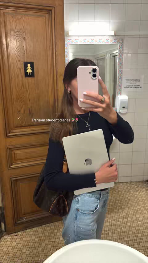 A young woman in a bathroom, taking a mirror selfie. She is holding a laptop and a phone. The background shows a wooden door and white tile.