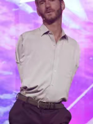 A medium shot shows a well-dressed man, likely of Middle Eastern descent, standing on a stage. He has short, neatly styled dark hair, a well-groomed dark beard, and is wearing a light-colored button-down dress shirt and dark pants. Interestingly, he has no arms visible. The background is a blurry gradient of pink and purple, suggesting a stage setting with focused lighting. The man appears to be speaking or presenting, with a serious expression.