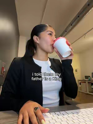 A woman is seated at a desk, drinking from a white and red cup. The text overlay reads, 'i told them my father is a King'. She wears a black jacket, a white top, and a ring on her left hand. The image has a soft, natural lighting.