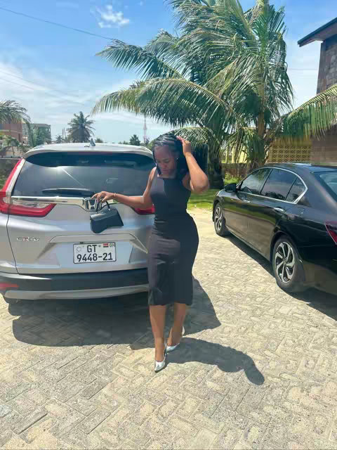 A woman in a black dress stands beside a silver car, holding a small black purse, in front of a background of a leafy tree and blue sky. The lighting appears sunny.