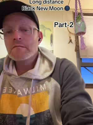 A man is centered in the frame, wearing a baseball cap, glasses, and a grey hoodie. Text overlays read 'Long distance Black New Moon Part-2'. In the background, a hanging plant is visible, and there is warm-toned interior decor.