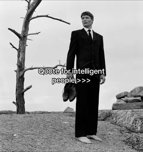 A black and white photo of a man in a suit holding shoes, standing on a rocky surface with a barren tree behind him and a cloudy sky.