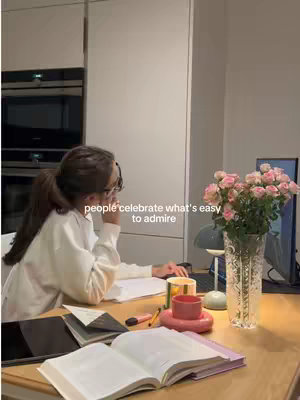 A person, likely a student, is working at a desk. The scene is indoors, probably in a kitchen or dining area, and the lighting is warm. The desk is cluttered with books, papers, and a laptop. In the background, a flower arrangement in a vase is visible, adding a touch of color and decoration to the scene.