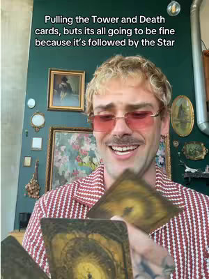 A man is holding tarot cards and looking towards the camera with a smile. Overlaying text on top of the image, a decorated wall is visible in the background.