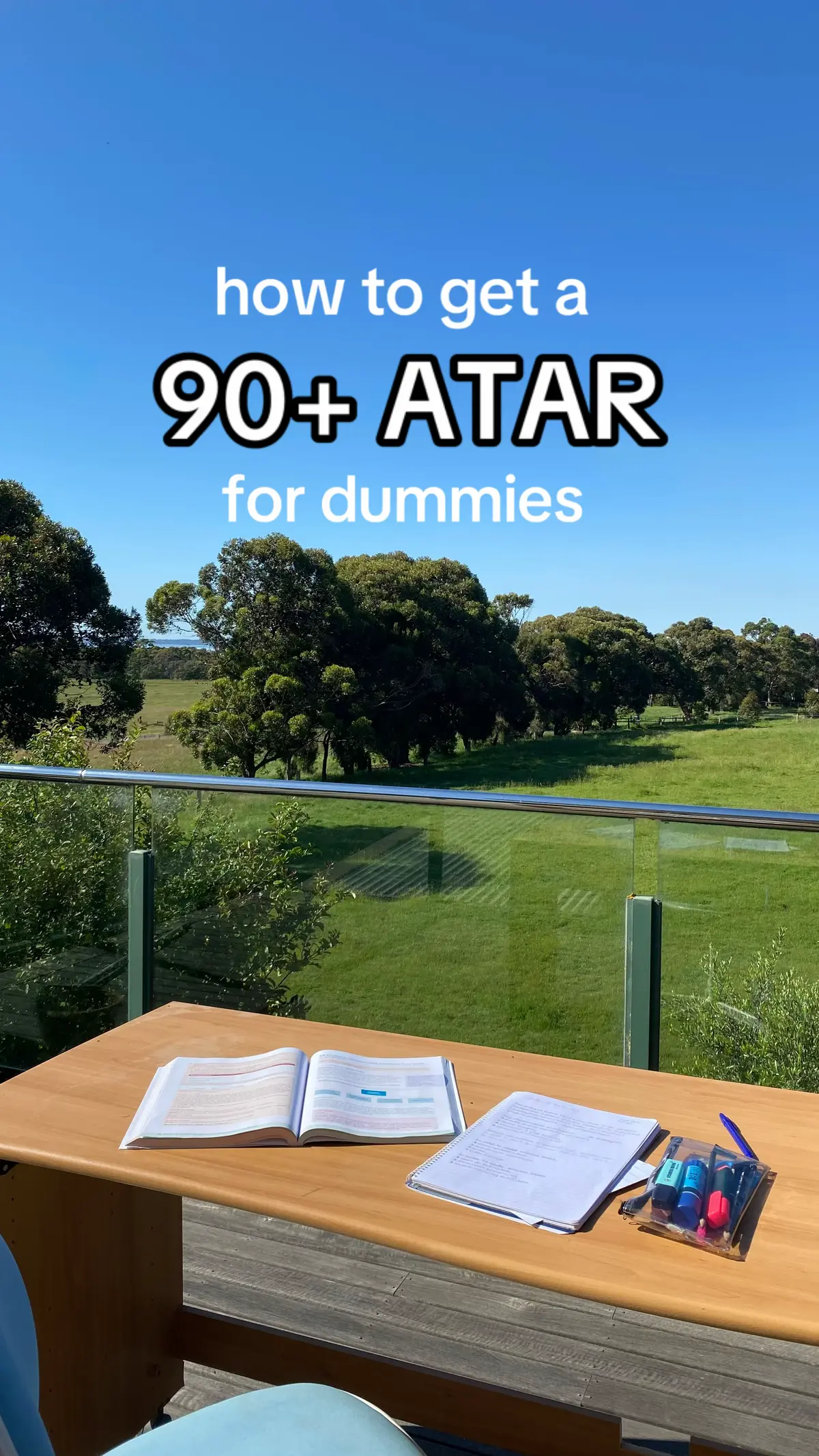 The cover image shows a clear blue sky above a lush green landscape. The foreground includes a wooden table with an open book, papers with writing, and a clear case of colored markers.  Above the table the title says 'how to get a 90+ ATAR for dummies'.