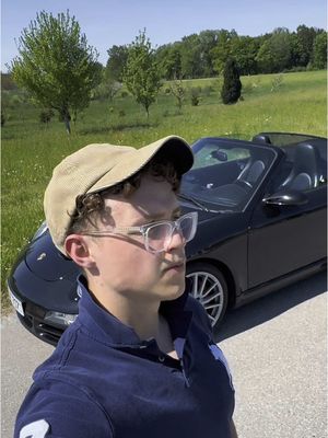 A selfie shows a young man in a tan corduroy cap and clear-frame glasses, posing next to a black convertible car on a paved road. The background features green fields, trees, and a bright blue sky.