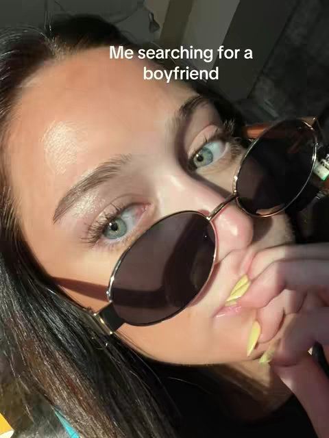 A close-up selfie of a young woman with long black hair. She's wearing round sunglasses, and her hand is near her mouth, with yellow nail polish visible. The background is dark and out of focus.