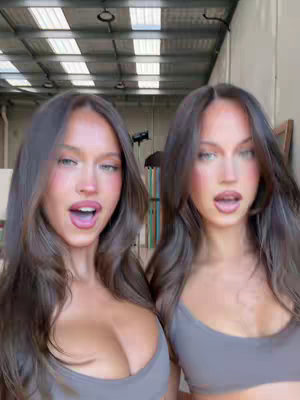 The cover image features two young women, both with long, dark hair and similar facial features, positioned side-by-side. They wear matching gray tops and are looking at the camera with open mouths, creating a sense of mirroring or duplication. The background is a high ceiling with industrial-style lighting, adding an urban and somewhat raw aesthetic to the shot.