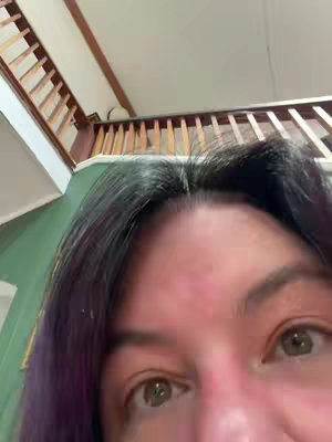 A close-up shot showing a person's eyes and the top of their head. The eyes are brown and looking upwards. The person has dark hair with purple highlights. The background shows a wooden staircase and a green wall.