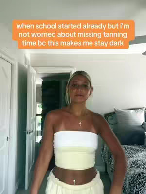A woman stands in a brightly lit room, likely a home interior, with white doors and neutral-colored walls visible in the background. She's wearing a white and yellow outfit and has blonde hair. Text overlaid on the image states, "when school started already but I'm not worried about missing tanning time bc this makes me stay dark."