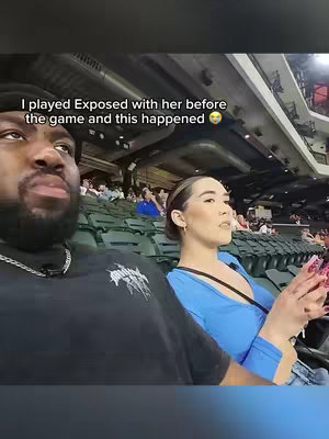 The image captures a couple seated in a stadium, presumably watching an event. The man, on the left, has dark skin, a beard, and wears a black shirt with a graphic, and a silver chain. He looks slightly up and to his right, his expression serious. To his right, the woman, who appears to be Asian, wears a blue top and has long, dark hair pulled back. She's holding a phone with pink phone case and looking slightly away, her expression contemplative. The background features green stadium seats filled with other spectators, with lights on. The overall composition suggests a casual setting.