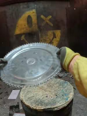 A person wearing gloves holds a large, metallic, circular object with a toothed edge over a stack of items. Behind them is a wall with a painted yellow and black design.