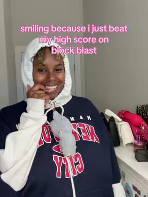 A woman with dark skin and curly hair is smiling, wearing a white hoodie with the hood up and a dark blue sweatshirt underneath that says 'MAN NEW YORK CITY' in large red letters. A small, gray and white bird is perched on her left sleeve. The background is a light-colored wall with some items on a shelf behind her.