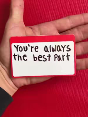 A hand holds a small white card with a red border against a textured red background. The card contains black handwritten text. The background is a solid red color.