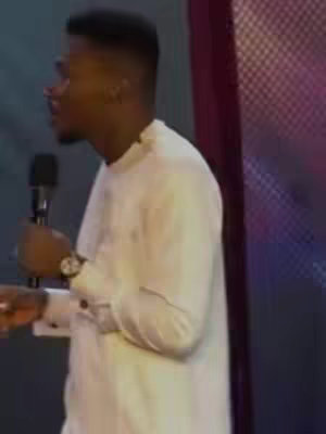 A man in a white shirt speaking into a microphone. He is standing in front of a blurred background with purple and dark sections.