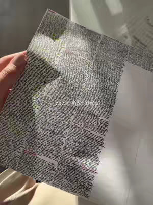 The image shows a hand holding a piece of paper covered in dense, small text. The paper is slightly angled, revealing a plain white surface behind it. The lighting is soft and indirect.