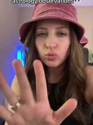 A woman with long brown hair is shown from the chest up, wearing a pink bucket hat and a black top. She's holding out her hand towards the camera, with fingers outstretched. There is text overlaid at the top of the frame with a playful font.