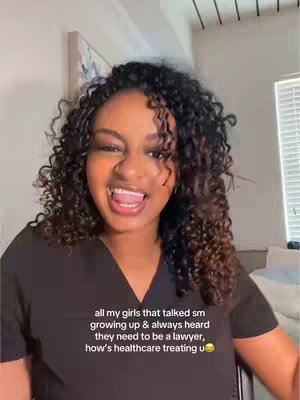 A smiling woman with curly hair is centered in the frame, looking directly at the camera with her mouth open. She is wearing a dark v-neck top. Text overlays are at the bottom of the frame, and the background includes a wall and a glimpse of an adjacent room.