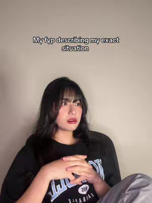 A woman with long, dark hair and a black graphic t-shirt looks at something off-camera with a concerned expression. The overlay text reads 'My fyp describing my exact situation'. The background is a plain wall.