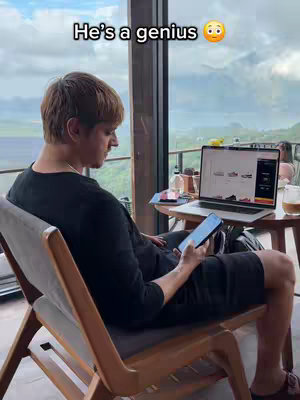 A man is seated in a chair with a laptop open on a small table to his side, holding a smartphone. The background hints at an outdoor setting with visible greenery and a clear sky.