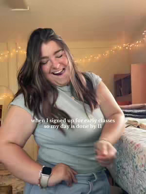 A woman with long brown hair is smiling and looking down. She is wearing a light blue t-shirt and a smartwatch with a light-colored strap and a silver watch face. Background is out of focus but contains some string lights.