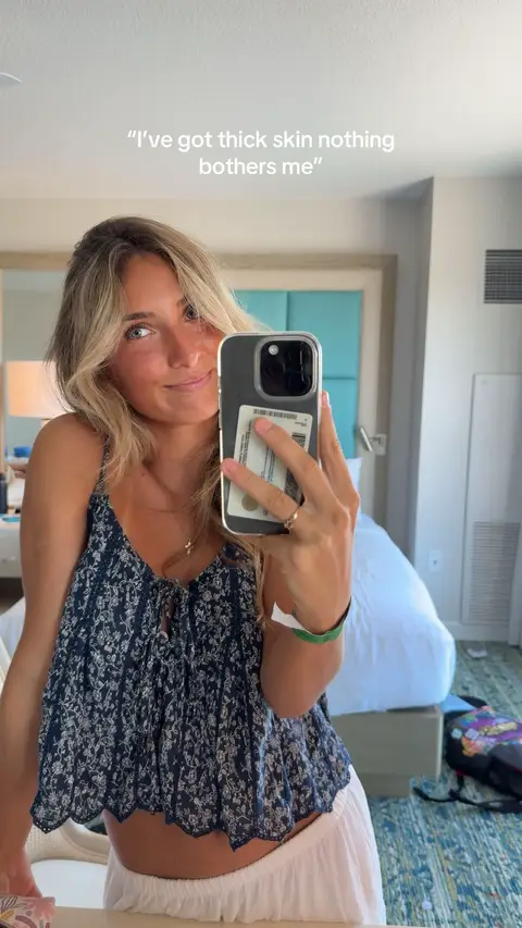A woman is taking a mirror selfie with her phone, showing a hotel room setting. She has light blonde hair and is wearing a patterned top. The text overlay says, "I've got thick skin nothing bothers me". The background includes a bed and other furniture.