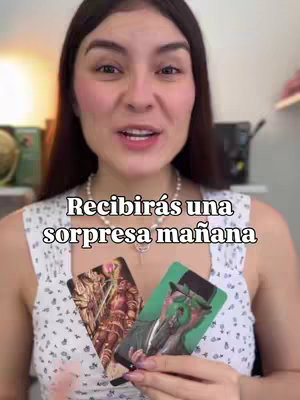 The image shows a woman in a close-up shot, smiling and looking at the camera. She is holding two tarot cards, with text overlaid on the image. The background is slightly blurred, with a glimpse of a room setting. The woman has dark hair and is wearing a white top, and a necklace.