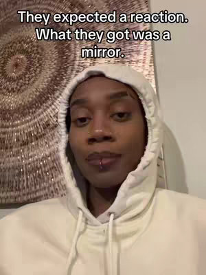 A person wearing a white hoodie on a medium shot. The background shows a brown circular design, and text at the top. The person has a lip piercing.
