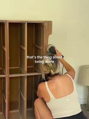 A woman is sanding a wooden cabinet, seen from behind and slightly from the side. She is in the middle of the frame, wearing a white tank top and dark shorts. Her hair is blonde and tied up. The upper section of a wooden cabinet is to the left of her, the cabinet is half-filled with shelves. In the background to the right, there is a wall in light-yellow, and towards the right, a lamp. The text overlayed is towards the center left.