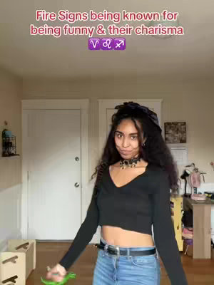 A young woman with long, dark hair and a black top stands in a room with a light-colored door and walls. Text overlays the image, with the main text at the top and zodiac symbols in purple. She is smiling with a slight frown. She poses with a green dinosaur toy in her hand.