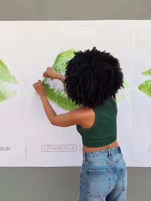 A person with dark curly hair is standing in front of a white wall with large green images of what appear to be lime wedges or leaf shapes. They are holding a white piece of paper and appear to be adding it to the wall. The person is  wearing a dark green tank top and light-wash blue jeans. The colors are bright and saturated. To the left of the woman, the word "SKIN" is visible, and below her are the words "CHIPOTLE", separated by a gray rectangle. The person is standing from a medium-low angle.