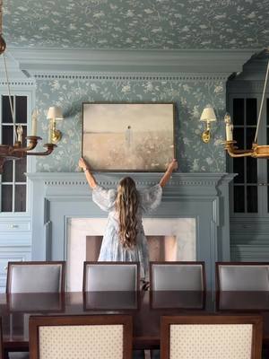 A woman with long blonde hair is holding a framed painting above a blue fireplace and mantel. The room features blue patterned wallpaper and a long, dark wood dining table with chairs.