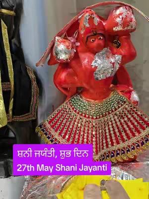 A vibrant image featuring a red idol figure, likely a Hindu deity, adorned with decorative elements. The figure has a detailed skirt with red and gold patterns, and foil accents on the head. Two texts in Punjabi and English are on a purple box over a part of the image.