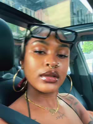 A close-up shot of a person inside a car. The person, who appears to be Black with dark skin, looks directly at the camera with a slightly raised eyebrow. They are wearing large gold hoop earrings, a gold chain necklace, and a nose ring. Black-rimmed glasses are perched on their head. The background remains slightly blurred, focusing on the person.