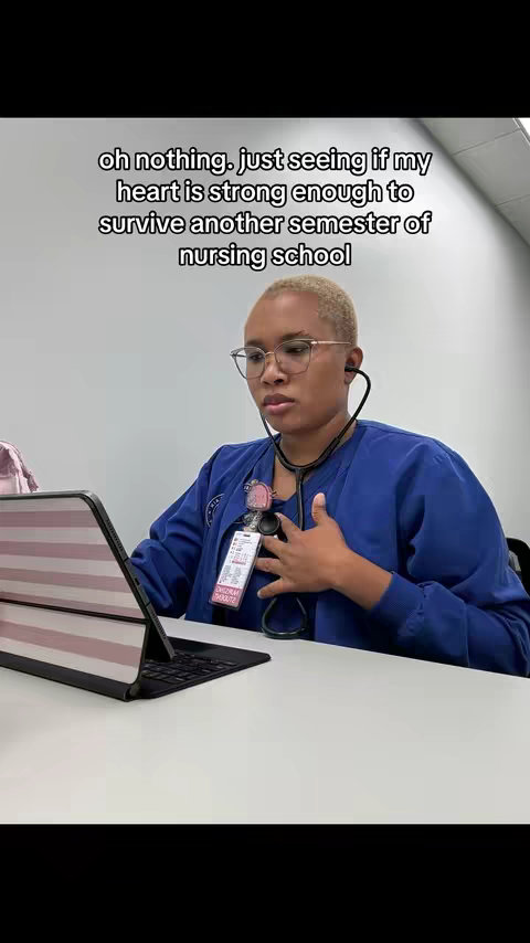 A woman in a blue scrub top, wearing glasses and a stethoscope, sits at a desk with a laptop open. She touches her chest.