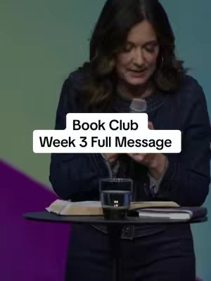 A woman with dark hair, wearing a dark denim jacket, stands behind a podium. She is looking down, likely reading from notes or a book on the podium. In front of her is a glass of water. A text overlay at the top reads: Book Club Week 3 Full Message.