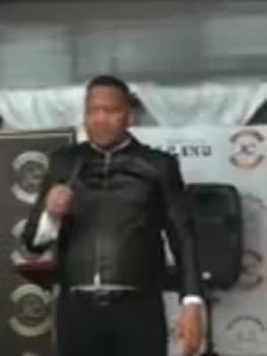 The cover image features a man standing in front of a backdrop, holding a microphone. He is wearing a black jacket over a white shirt, suggesting a formal or professional setting. The background includes banners with indistinct text and logos. The image is of average quality, suggesting it may have been taken from a video.