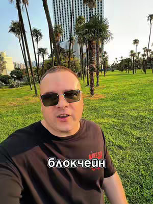A man wearing sunglasses and a black t-shirt stands in front of a green, grassy area. Behind him are tall palm trees and a modern-looking building.